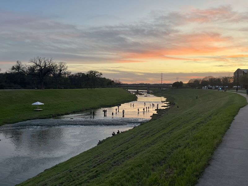 The Clear Fork of the Trinity at dusk: the paved trail running along the bank, anglers standing out in the shallows, sunset over the bridge.
